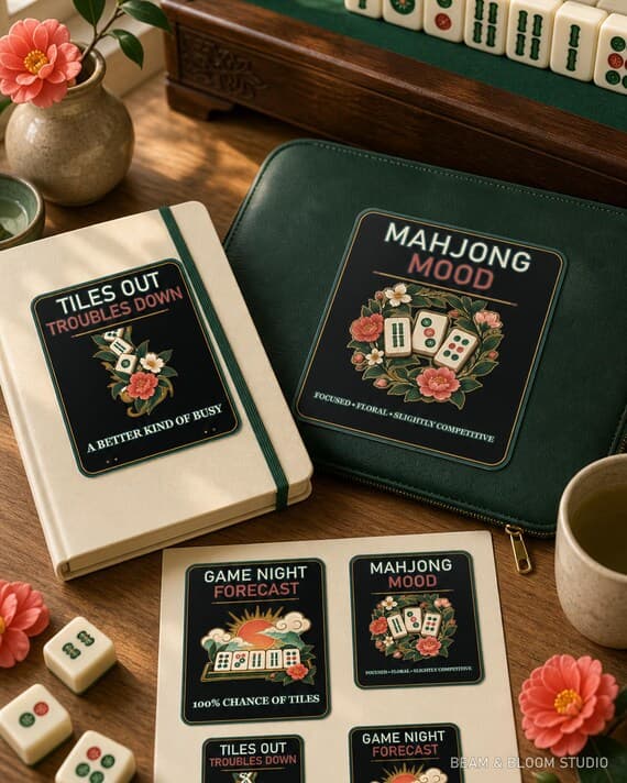 Mahjong game-night designs shown as coordinated stickers on a notebook and laptop sleeve in a warm maker scene.